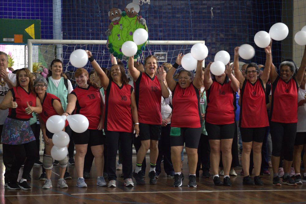 Na imagem, há um grupo mulheres, de diferentes idades, usando coletes na cor vermelha e segurando bexigas brancas com os braços erguidos, todas sorrindo e vibrando. Elas estão em uma quadra poliesportiva com uma trave e redes ao fundo